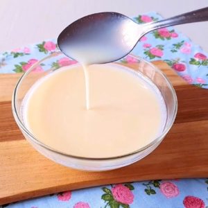 Tigela de vidro com calda cremosa de leite em pó para cobertura branca de bolo, sendo colherada para demonstrar a consistência lisa e fluida.