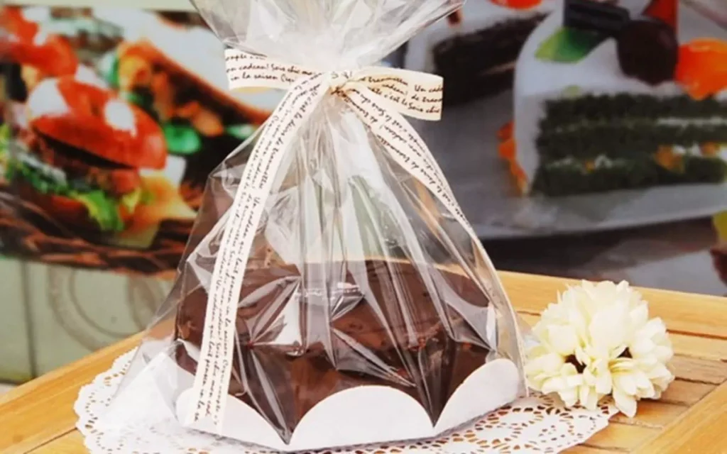 Bolo ou doce embalado elegantemente em plástico transparente com laço de fita, em cima de um doily e base branca, destacando a importância da embalagem na confeitaria.