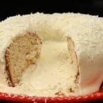 Bolo branco com cobertura de leite ninho cremosa e aerada, decorado com raspas de chocolate branco. Uma fatia cortada revela o recheio macio do bolo e a textura sedosa da cobertura.