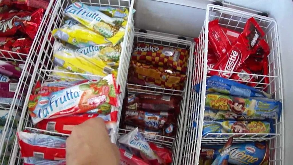 Freezer horizontal aberto com cestos metálicos cheios de picolés e sorvetes embalados (La Frutta, Nestlé), demonstrando a organização interna essencial para otimizar o estoque e a venda por impulso em sorveterias.