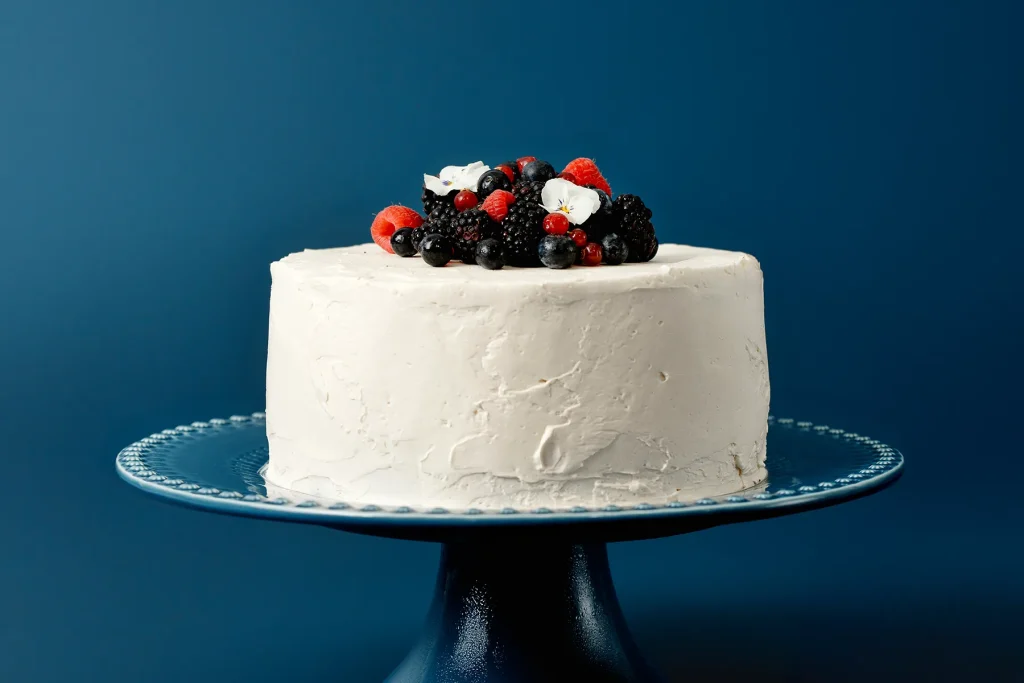 Bolo de festa com cobertura branca de buttercream de merengue suíço perfeitamente alisada, decorado com um mix de frutas vermelhas e mirtilos no topo. A cobertura firme é ideal para bolos de aniversário.