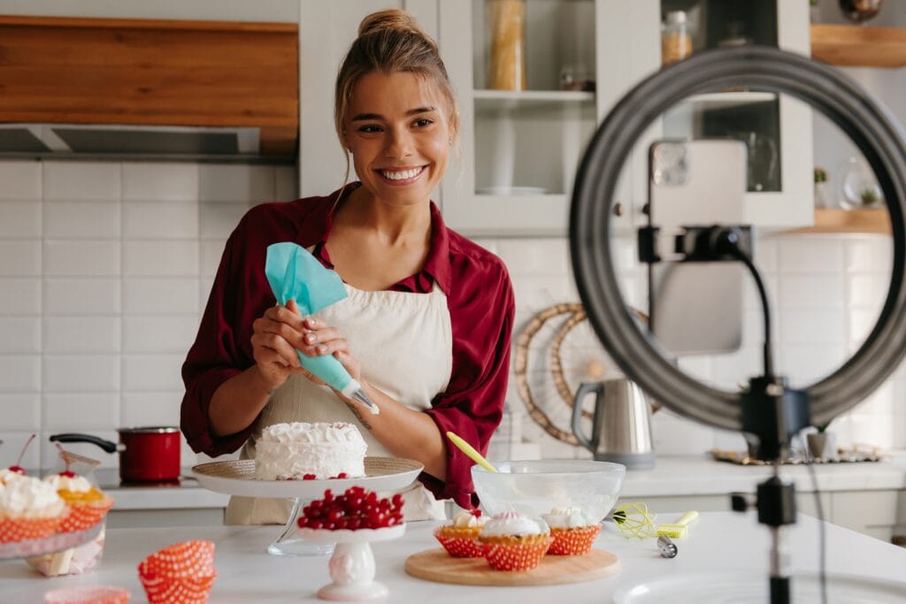 Mulher sorridente gravando um vídeo de decoração de mini bolo com saco de confeitar em sua cozinha, utilizando um celular e um ring light para marketing de conteúdo.