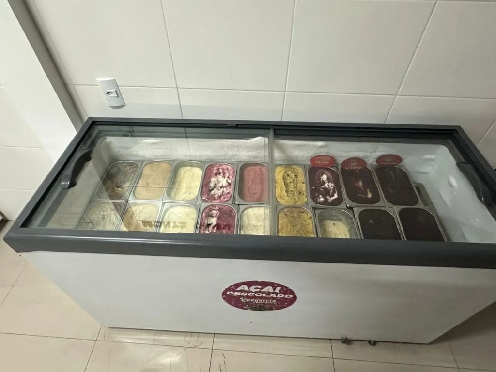 Visão superior de um freezer horizontal com tampa de vidro, exibindo 18 cubetas de sorvete e açaí de diferentes sabores perfeitamente organizadas e conservadas.