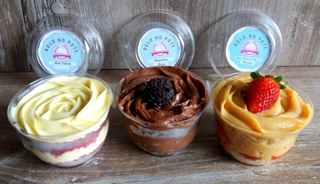 Três Bolos no Pote Gourmet (Red Velvet, Brigadeiro Belga e Doce de Leite) com apresentação premium, tampas e rótulos. O Apelo Visual ideal para atrair clientes em Feiras de Natal e eventos, posicionando o produto como Produto-Estrela.