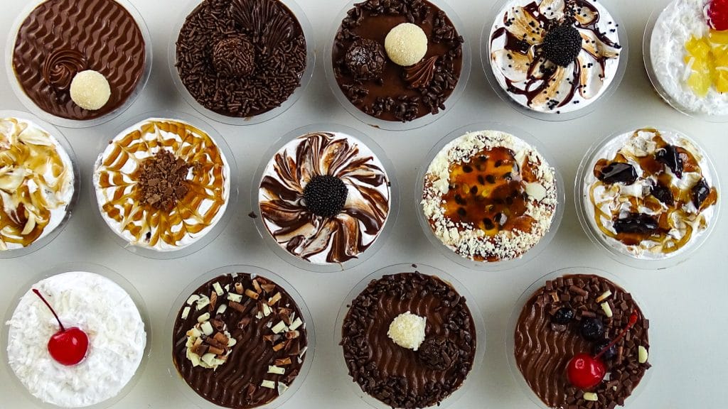 Amostra de diversos Bolos no Pote de sabores variados (chocolate, maracujá, caramelo, coco), prontos para a venda. A imagem demonstra o potencial de Escala de Produção viabilizado pelo Congelamento e o aumento da Validade do produto.