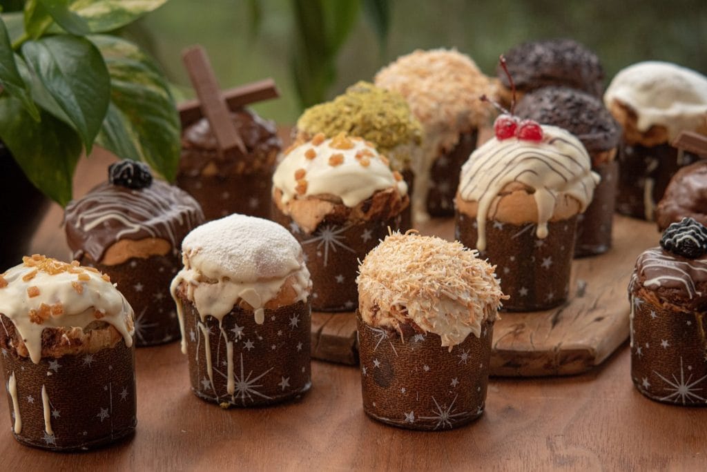 Mesa com sortimento de panetones gourmet de diversos sabores (pistache, chocolate, coco), cada um com embalagem e decoração diferenciada. O Menu Gourmet é a principal estratégia de branding para justificar o preço e maximizar o lucro.