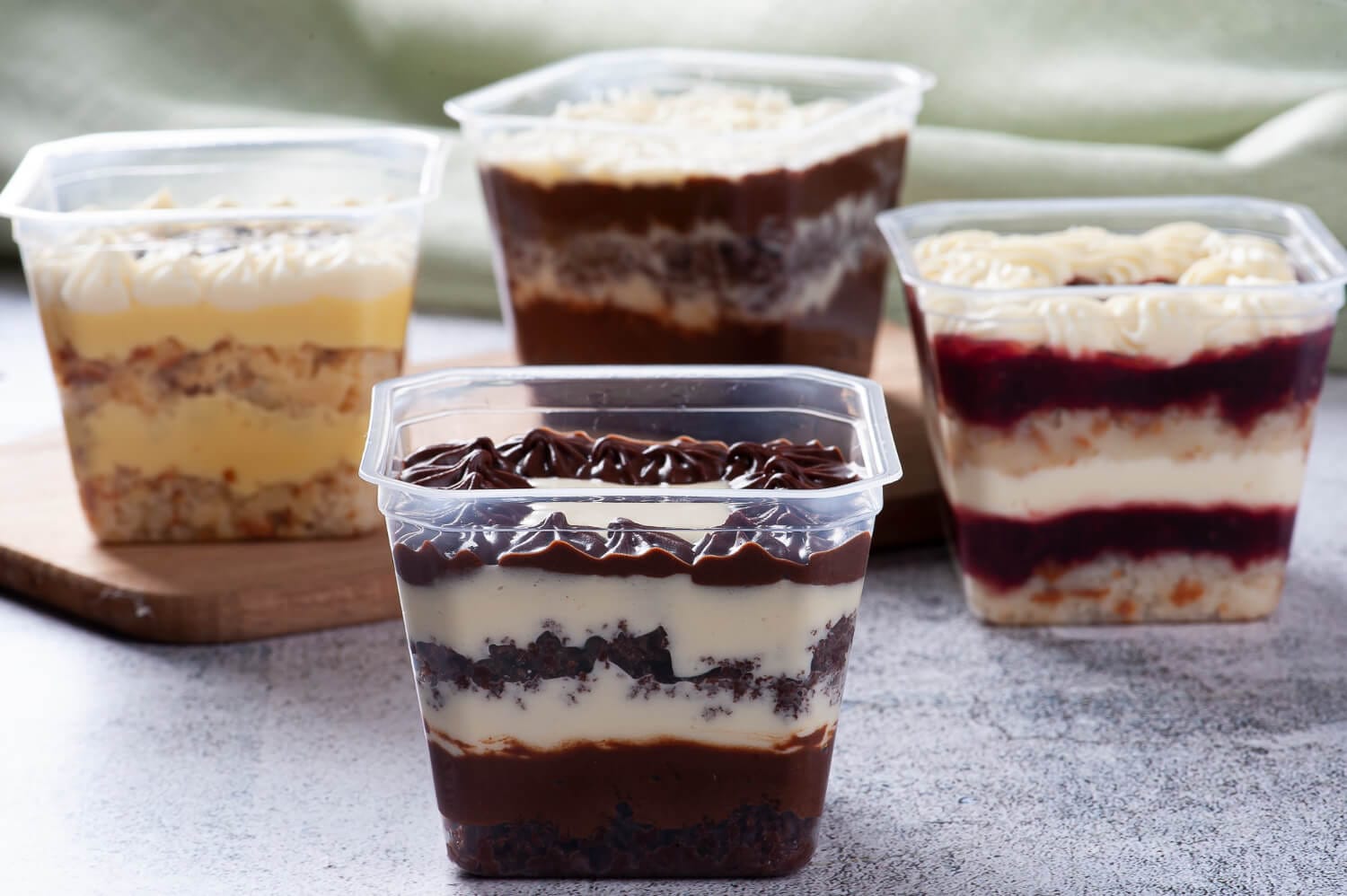 Quatro Bolos no Pote com camadas de recheios e massas contrastantes, embalados em potes transparentes de formato quadrado, ilustrando o alto padrão visual dos produtos Gourmet, Fitness e Veganos.