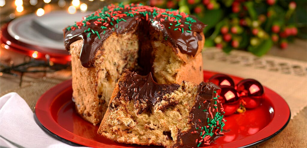 Chocotone decorado com cobertura de chocolate e granulado vermelho e verde, com uma fatia cortada. A imagem representa o produto final de um Natal Organizado, resultado de uma Estratégia de Antecipação e Logística de Tempo eficiente, garantindo Lucro e Profissionalismo.