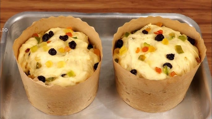 Duas massas de Panetone Caseiro com frutas cristalizadas e passas crescendo em formas de papel. A imagem representa o estágio de fermentação, que, combinado com o uso estratégico de Gordura, Gema e a técnica de Brine, garante a Umidade Duradoura e a maciez.
