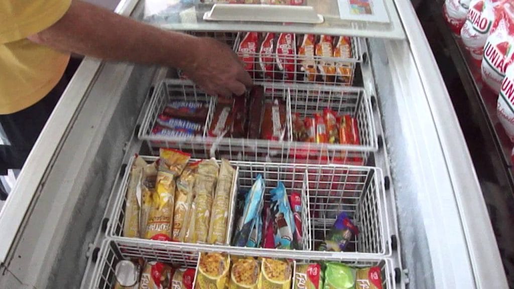 Visão interna de um freezer horizontal aberto, mostrando diversos sabores de picolés e potes de sorvete organizados em cestos metálicos, facilitando a aplicação do método FIFO e o controle de estoque.