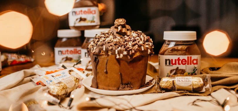 Panetone Gourmet de Nutella ricamente decorado em um Cenário de Luxo com embalagens originais. A foto de alto impacto serve como Gatilho de Venda por Impulso, construindo a Autoridade Visual da marca no Natal.