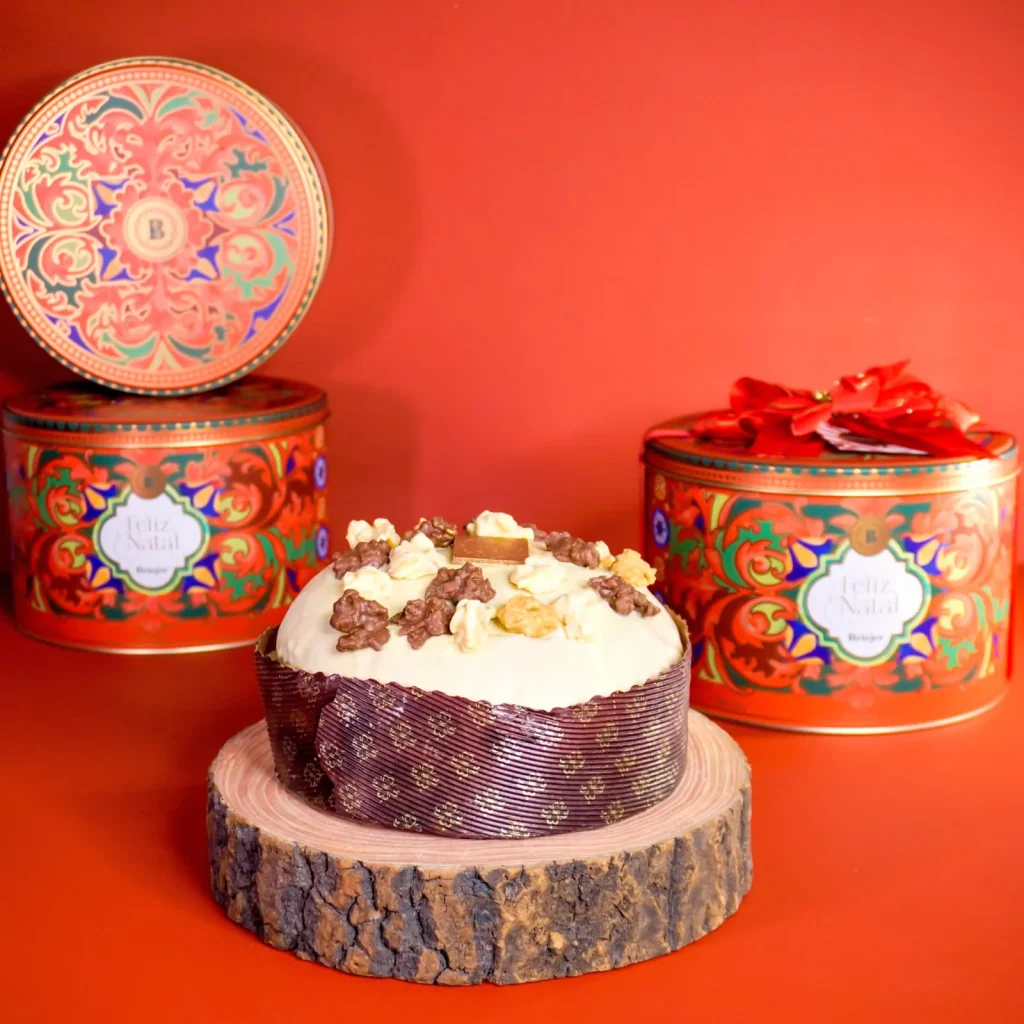 Panetone com cobertura branca e decoração em cima de um display de madeira rústica. Ao fundo, três Latas Estilizadas de Natal. A imagem, inspirada na marca Biscoitos Briejer, ilustra o conceito de Embalagem Premium (Lata) para o Chocotone na Lata, vendendo-o como um presente de luxo.
