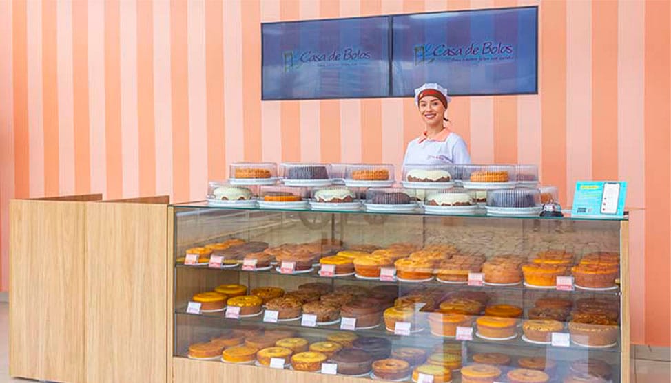 Loja de Confeitaria bem-sucedida com balcão cheio de bolos, ilustrando o alto Faturamento e a Autoridade em Vendas. O sucesso é resultado da Estratégia de Distribuição Multicanal (Delivery, Feiras e Consignação).