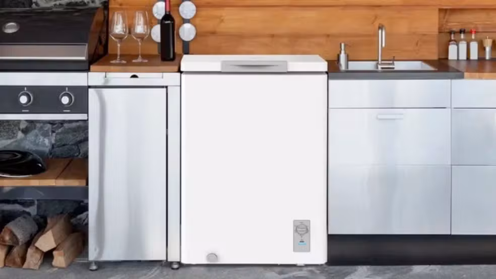 Um freezer horizontal branco compacto instalado entre uma bancada de aço inox com pia e um armário planejado em uma área gourmet com parede de madeira e churrasqueira ao lado.