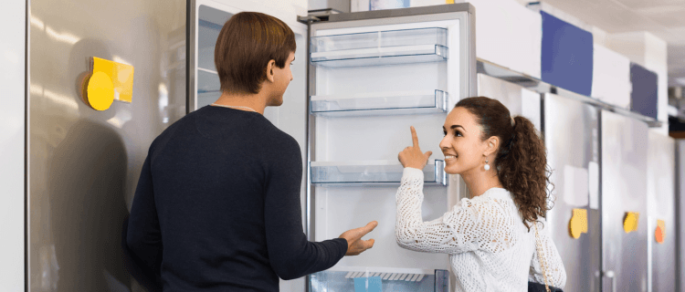 Um homem de costas e uma mulher sorrindo apontam para o interior de um refrigerador vertical aberto em uma loja, com várias outras geladeiras alinhadas ao fundo.