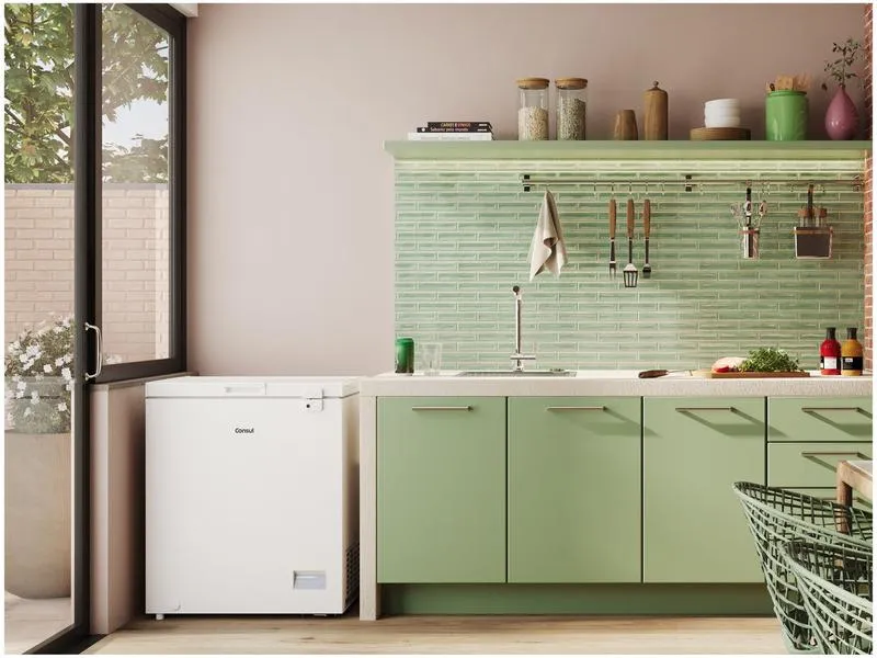 Um freezer horizontal branco da marca Consul posicionado em uma cozinha moderna com armários verdes, bancada clara e uma porta de vidro ao lado que permite a entrada de luz natural.
