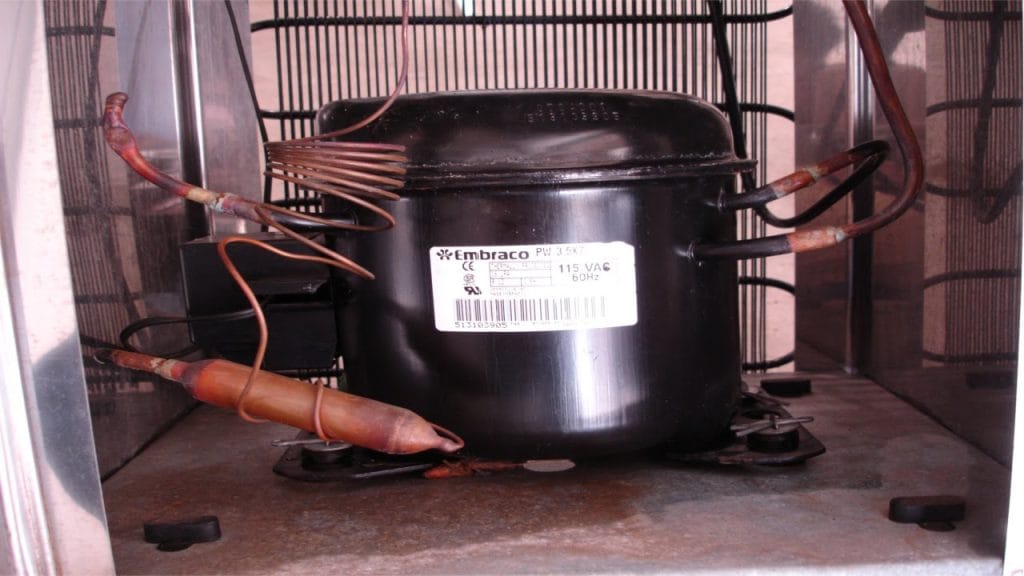 Vista aproximada de um motor compressor de freezer da marca Embraco, destacando a engenharia interna e os tubos de cobre que compõem o sistema de refrigeração e potência térmica.