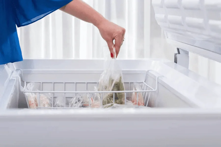 Mão de uma pessoa organizando sacos de alimentos congelados em um cesto aramado de freezer horizontal, representando o cuidado com a estabilidade térmica na confeitaria.