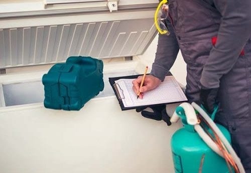 Técnico de manutenção usando uniforme cinza e óculos de proteção, fazendo anotações em uma prancheta ao lado de um freezer horizontal aberto e um cilindro de gás refrigerante verde.